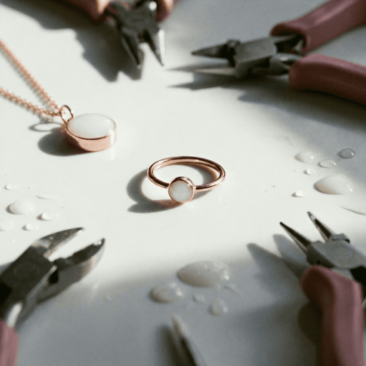 Sur un plan de travail, une bague lait maternel diy en métal rose avec pierre blanche, entourée de pinces et de gouttes de lait, à côté d’un pendentif assorti.