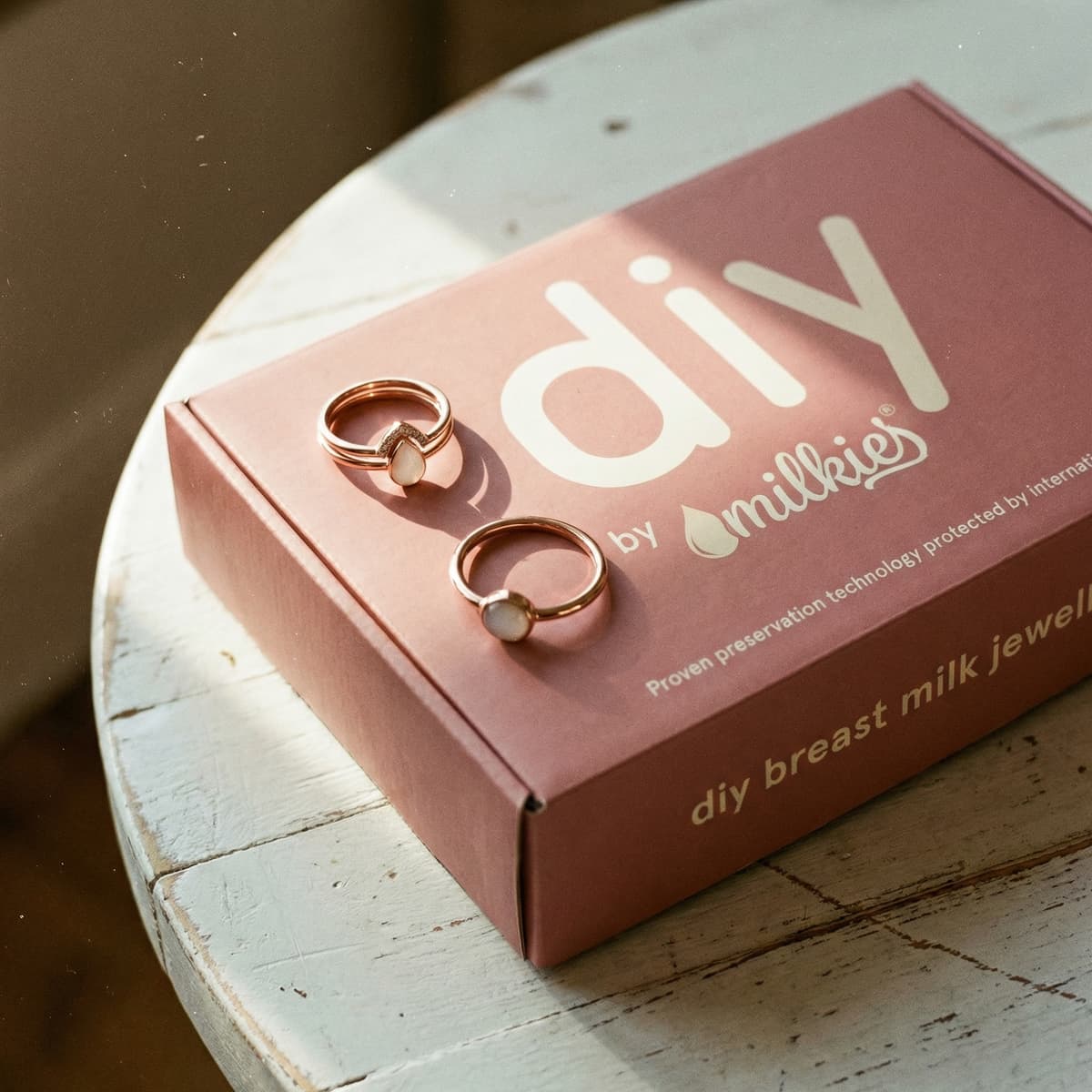 Deux bagues dorées en résine blanche, idéales comme bague souvenir allaitement DIY by MILKIES, posées sur une boîte rose du kit « diy breast milk jewel » sur une table en bois.