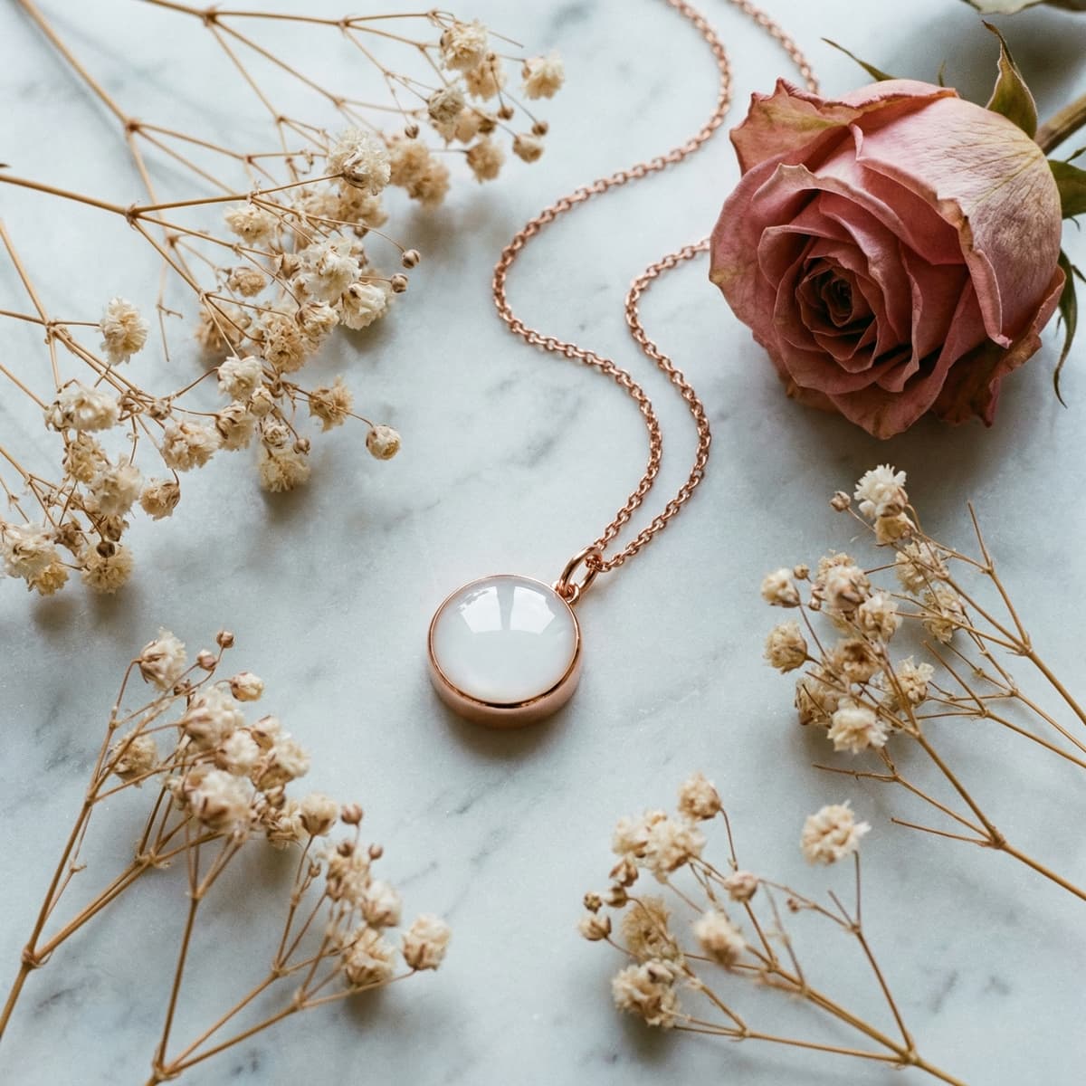 Collier délicat en chaîne dorée avec pendentif rond nacré, présenté comme un bijou souvenir lait, posé sur fond marbre avec une rose poudrée et des fleurs séchées.