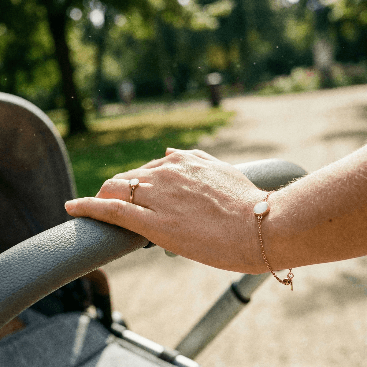 Main d’une maman tenant la poignée d’une poussette en promenade, portant une bague et un bracelet dorés avec pierre laiteuse, exemples de bijoux lait maternel en souvenir.