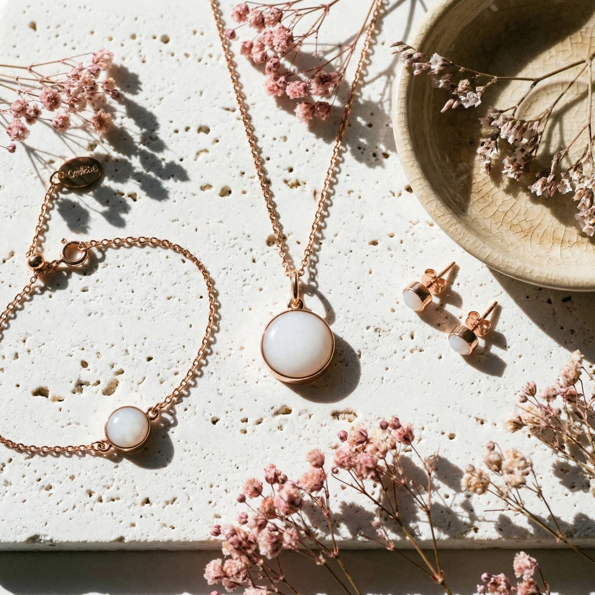 Bijoux en or rose avec pierre blanche laiteuse (collier pendentif, bracelet et boucles d’oreilles) sur surface en pierre, entourés de fleurs séchées, idée de cadeau fin d'allaitement délicat et symbolique.