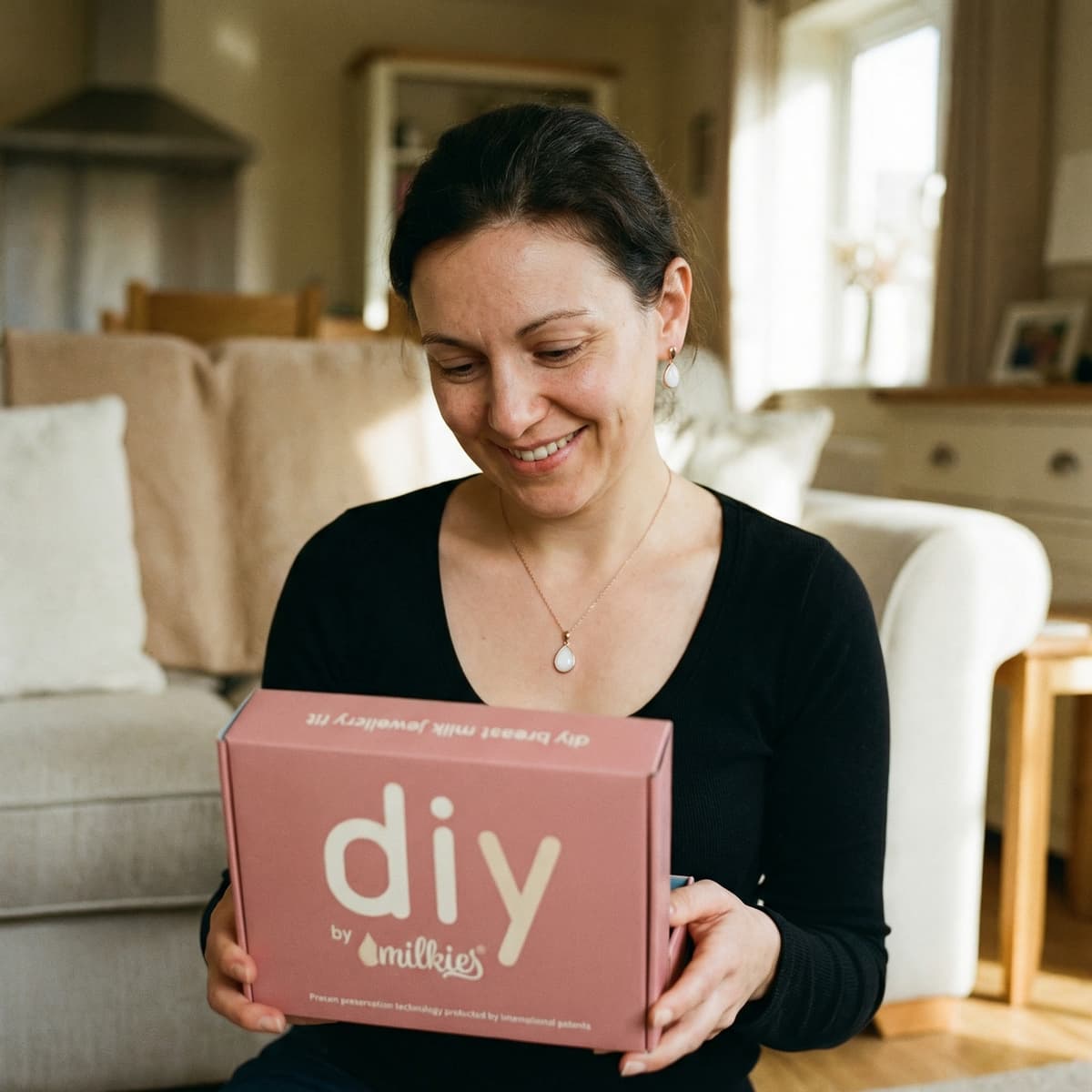 Jeune maman souriante à la maison découvrant la boîte du kit DIY by MILKIES, portant un collier goutte de lait et des boucles d’oreilles assorties, pour créer facilement un bijou souvenir en toute autonomie et confidentialité.