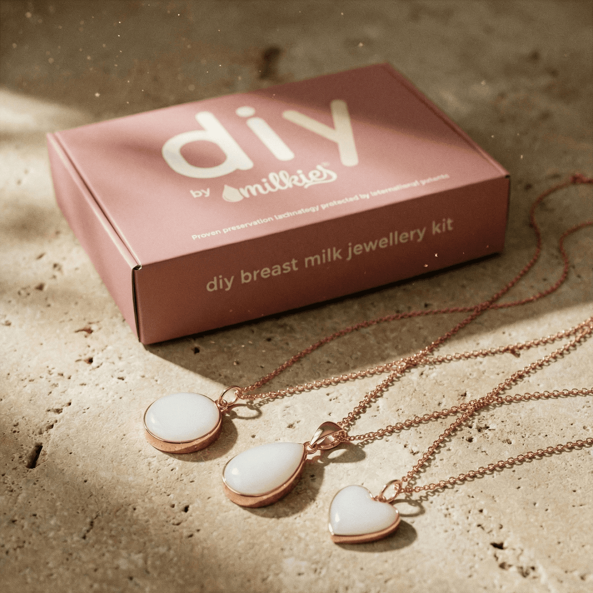 Kit DIY by MILKIES pour création collier lait maternel, avec boîte rose et trois pendentifs blancs (rond, goutte, cœur) sur chaînes rose gold posés sur une surface en pierre.