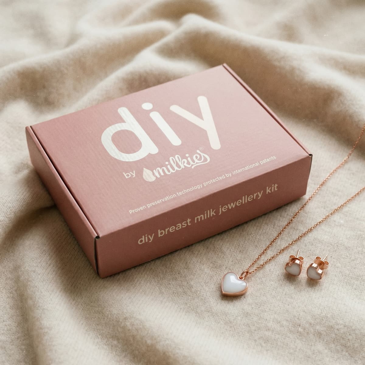 Boîte rose DIY by MILKIES, une idée cadeau jeune maman originale avec kit de bijoux au lait maternel et parure cœur rose doré (collier et boucles d’oreilles) sur tissu beige.