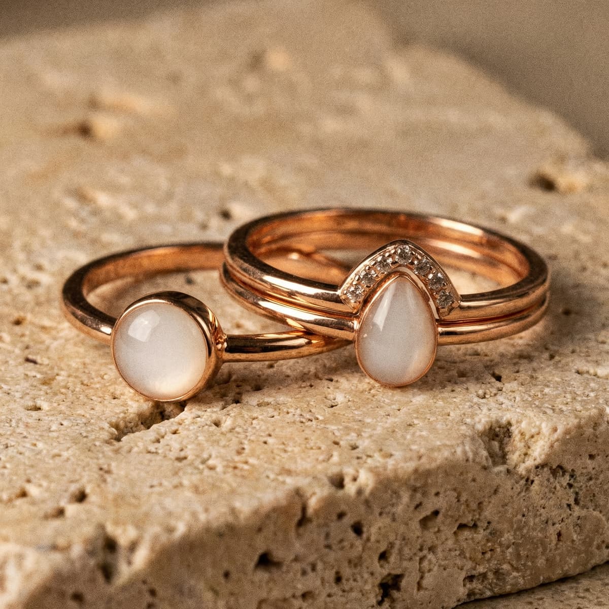 Deux bagues en métal doré rose avec pierres blanc laiteux et détails sertis, exemple de bijou souvenir réalisé avec un kit bague lait maternel, posées sur une surface en pierre.