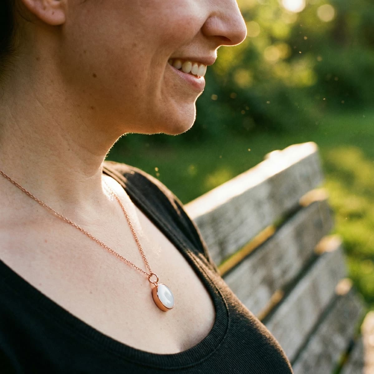 Portrait en plein air d’une femme souriante portant un pendentif ovale blanc sur chaîne dorée, réalisé avec un kit collier lait maternel pour créer un souvenir discret et élégant.