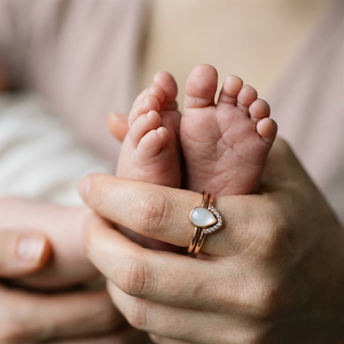 Gros plan d’une main de maman tenant les petits pieds de son bébé, portant une bague rose doré avec pierre en forme de goutte, symbole d’un souvenir allaitement intime et précieux.