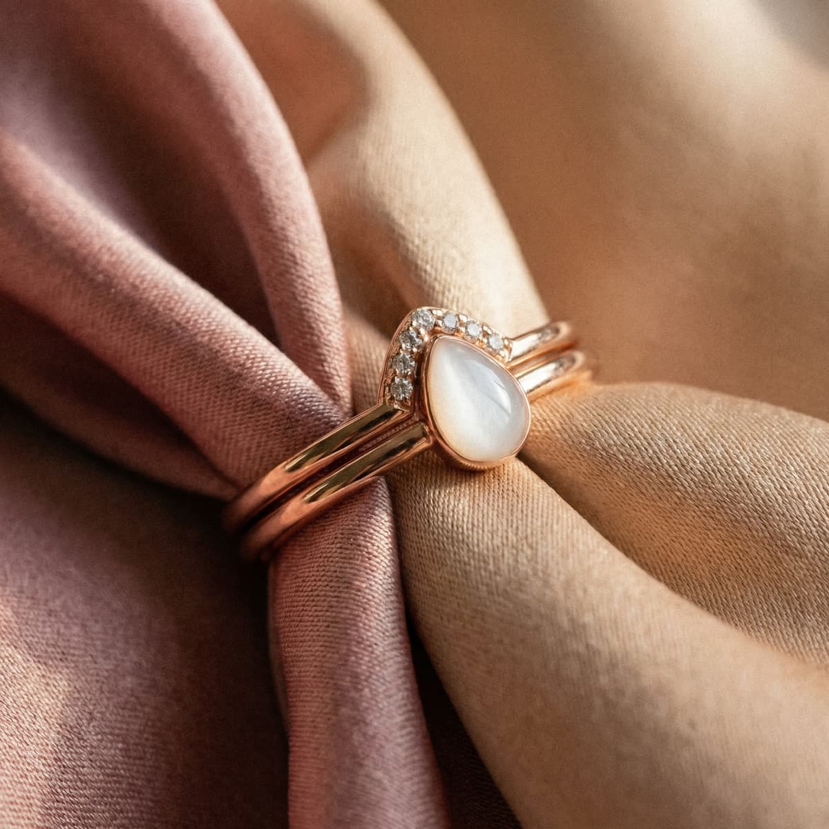 Bague en or rose avec pierre lactée en forme de goutte et pavage de petits diamants, idéale comme souvenir maternité raffiné, posée sur un tissu satiné rose et beige.