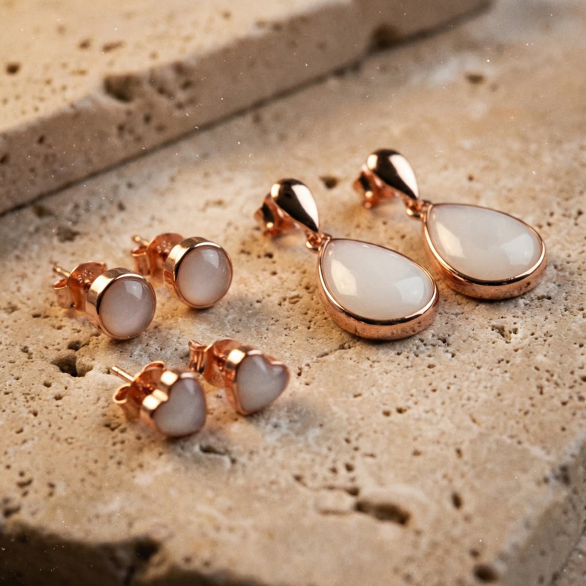 Assortiment de boucles d’oreilles en finition or rose avec pierre blanche laiteuse, modèles goutte, ronds et cœurs, inspirés des boucles d’oreilles allaitement diy, posés sur une surface en pierre texturée.