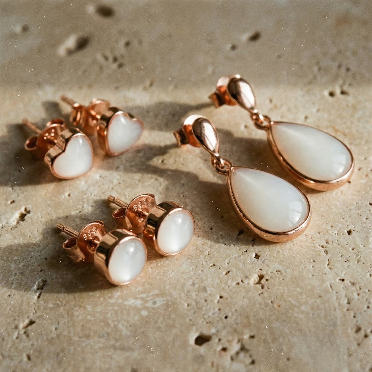 Ensemble élégant de boucles d’oreilles lait maternel en résine blanche, serties de métal doré rose, avec puces rondes et cœurs et pendants en forme de goutte, posées sur une surface en pierre.