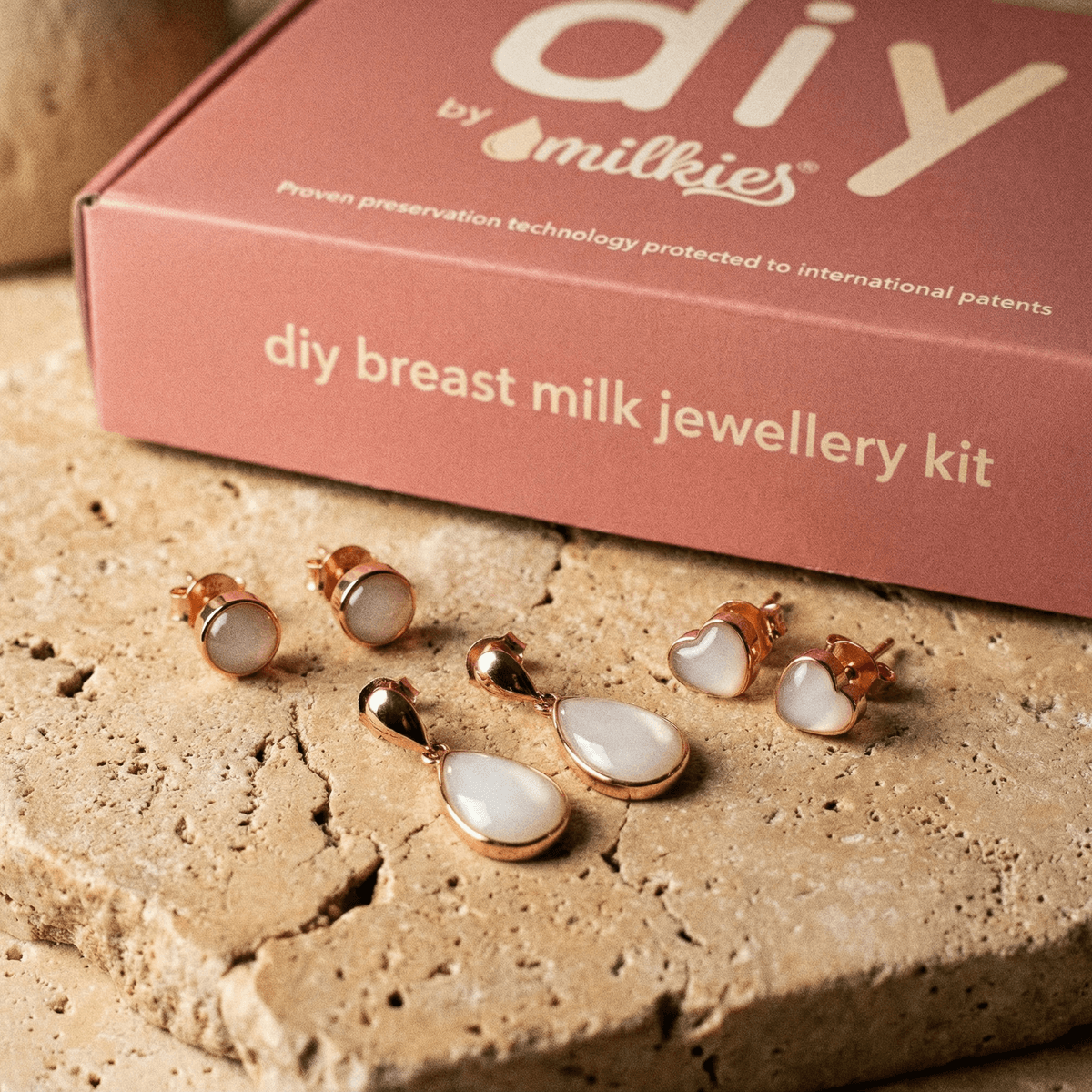 Présentation de boucles d'oreilles souvenir lait et pendentifs en résine blanche sertis rose gold devant une boîte de kit « DIY by Milkies » pour créer des bijoux au lait maternel.