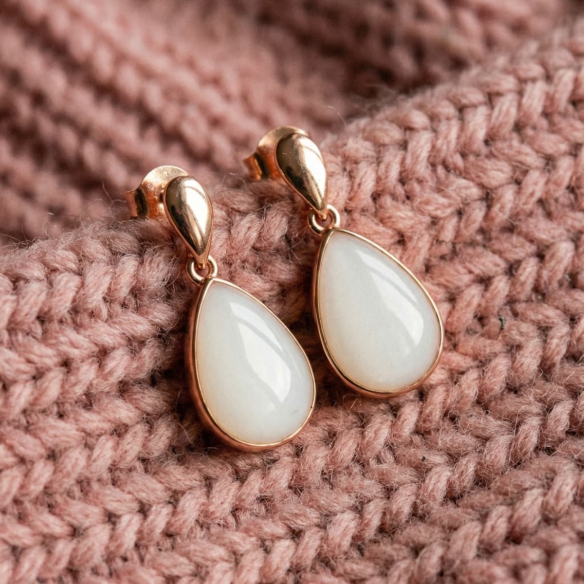 Boucles d'oreilles en forme de goutte avec cabochon blanc nacré et monture dorée, modèle de boucles d'oreilles souvenir lait, posées sur un tissu tricot rose poudré.
