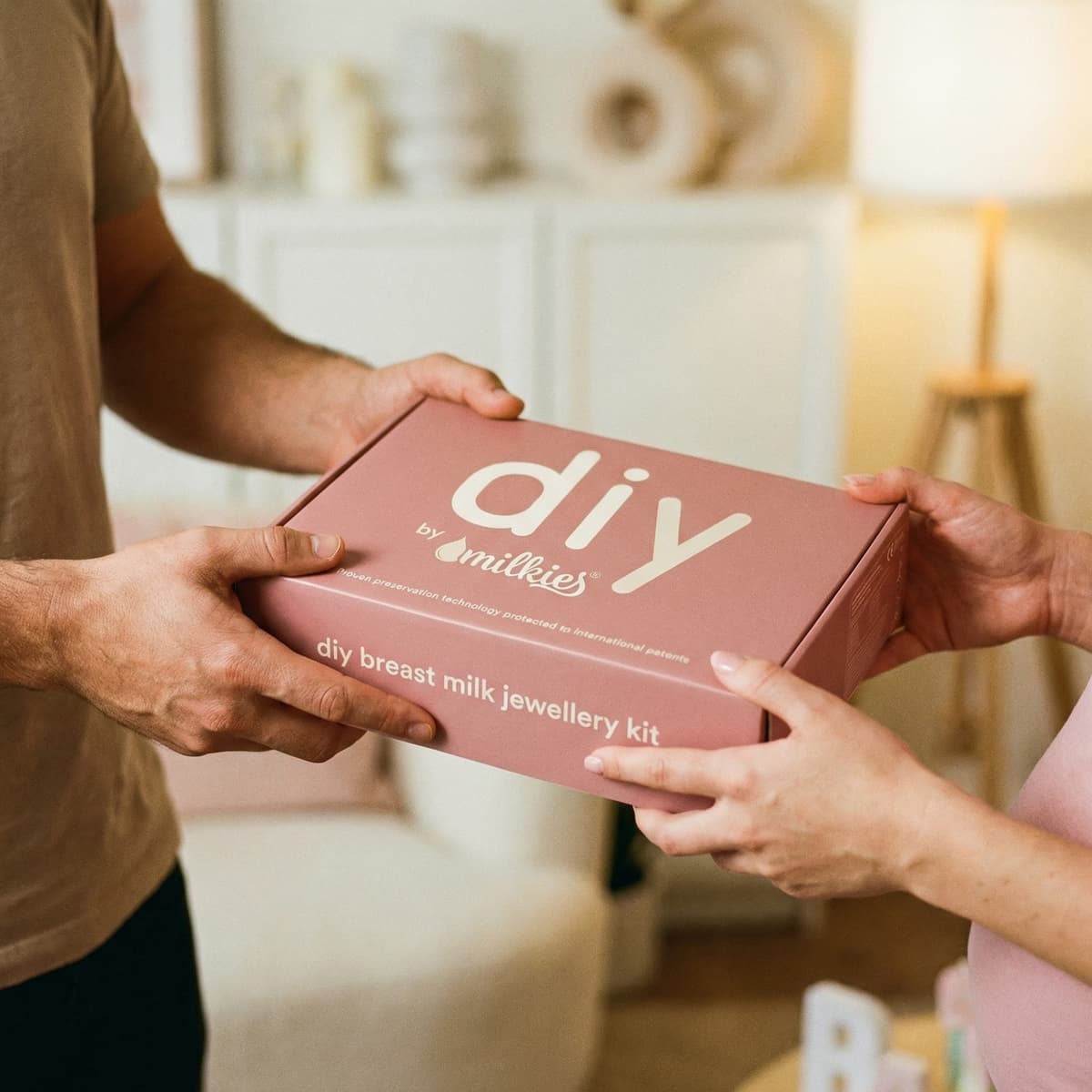 Deux personnes se passent une boîte rose DIY by MILKIES, kit de bijoux au lait maternel, idée de cadeau allaitement à réaliser chez soi.