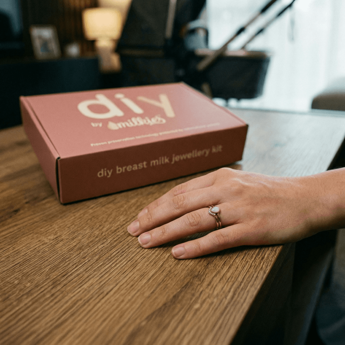 Main d’une maman sur une table en bois portant une bague souvenir, à côté d’un coffret DIY by MILKIES de création de bijoux au lait maternel, idée de cadeau naissance original maman.