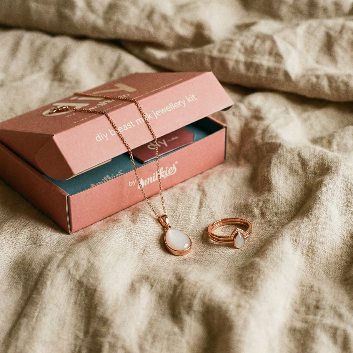 Idée de cadeau naissance pour sa femme : kit DIY by MILKIES dans une boîte rose, avec collier et bague finition or rose et pierre blanche posés sur tissu.