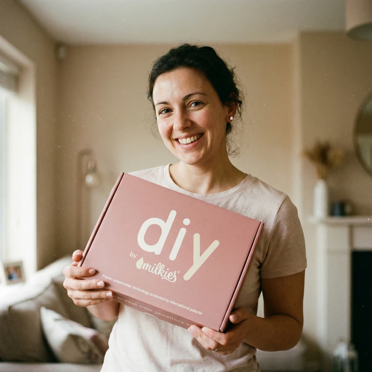 Jeune maman souriante tenant une boîte rose « DIY by Milkies », kit breveté pour fabriquer boucles d'oreilles lait maternel et autres bijoux souvenirs chez soi en toute simplicité, idéal pour créer un souvenir en toute intimité.