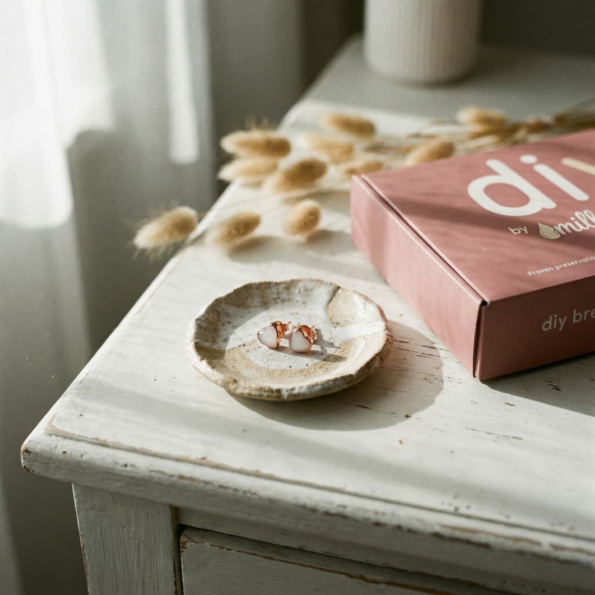Kit DIY by MILKIES posé sur une table blanche en bois, avec boîte rose et support doré présentant des boucles d’oreilles en résine blanche, montrant comment faire des boucles d'oreilles lait maternel à la maison.