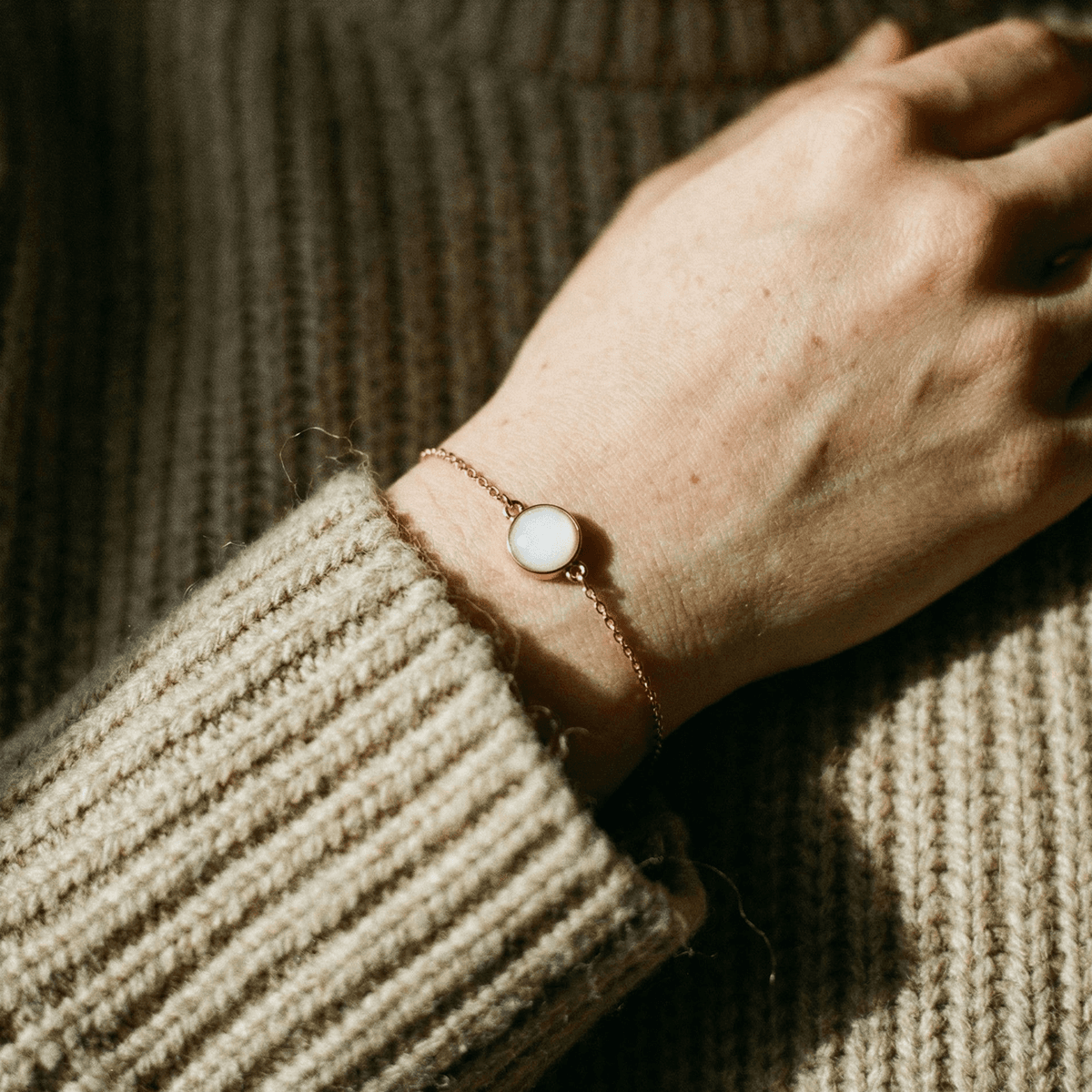 Poignet portant un bracelet fin doré avec cabochon blanc nacré sur un pull en maille beige, évoquant l’idée de faire un bracelet avec son lait dans une lumière douce.