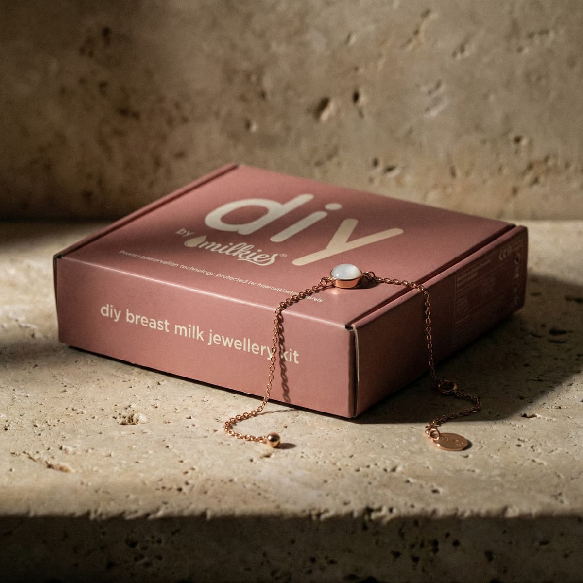 Kit DIY by MILKIES pour faire un bracelet avec son lait, présenté dans une boîte rose « diy breast milk jewellery kit » avec une chaîne rose doré et un médaillon blanc posés dessus.