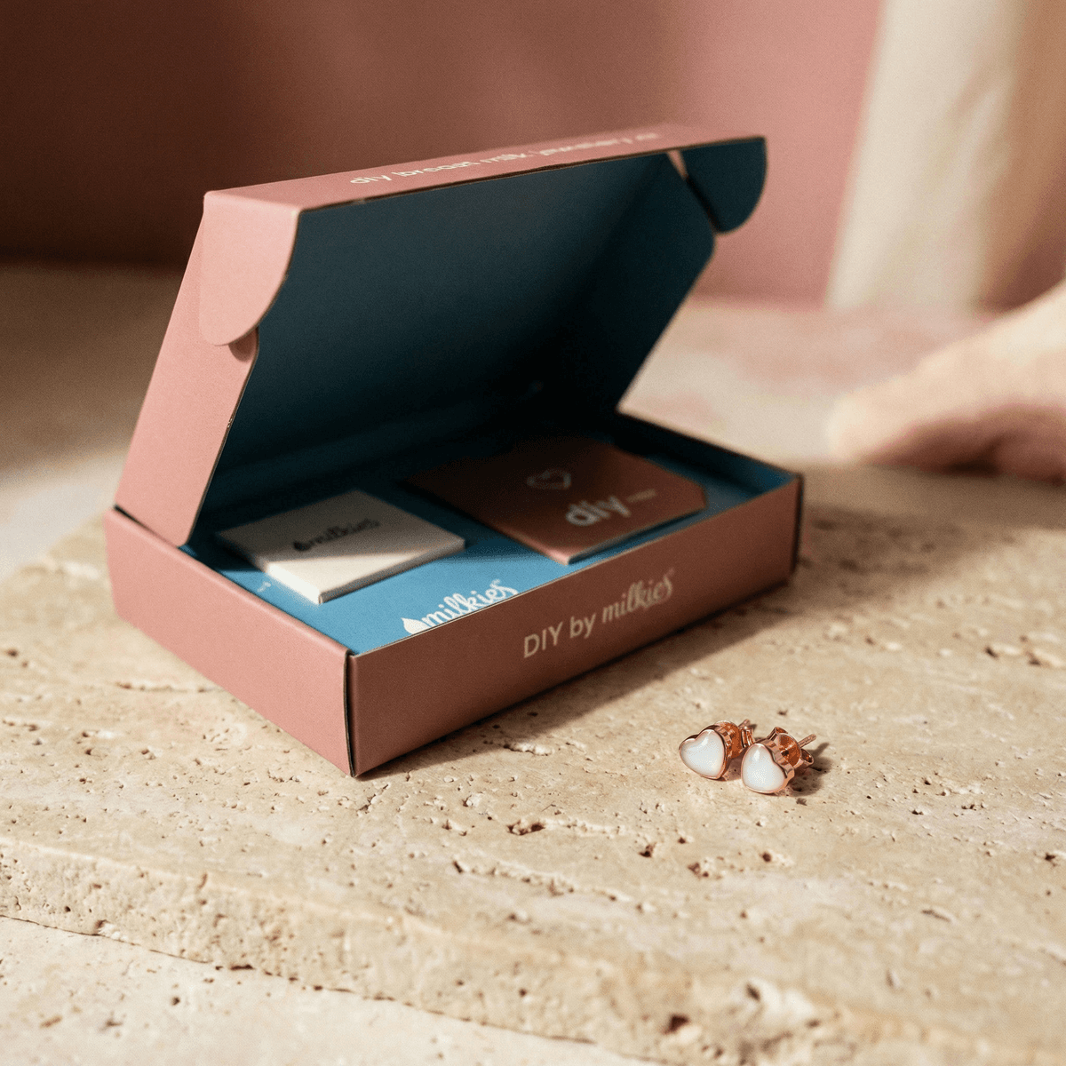 Coffret DIY by MILKIES ouvert sur une table, avec accessoires et supports pour créer des bijoux souvenirs, incluant une paire de boucles d’oreilles cœur rose doré du kit créatif boucles d'oreilles lait à réaliser chez soi.