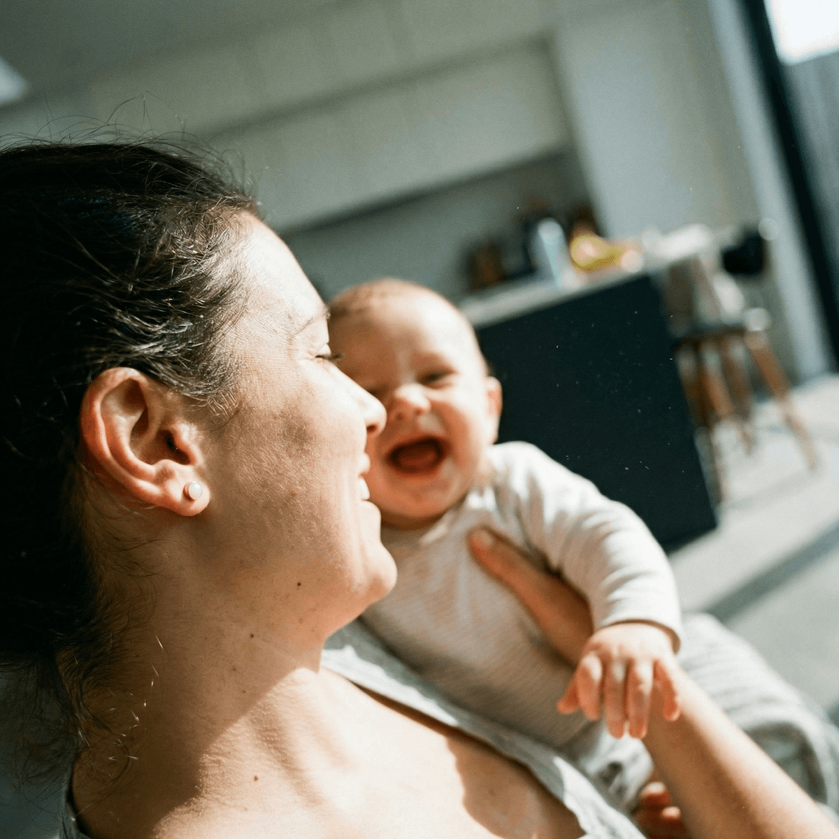 Jeune maman souriante tenant un bébé rieur à la lumière du matin, évoquant un souvenir de maternité à créer avec un kit créatif boucles d'oreilles lait.