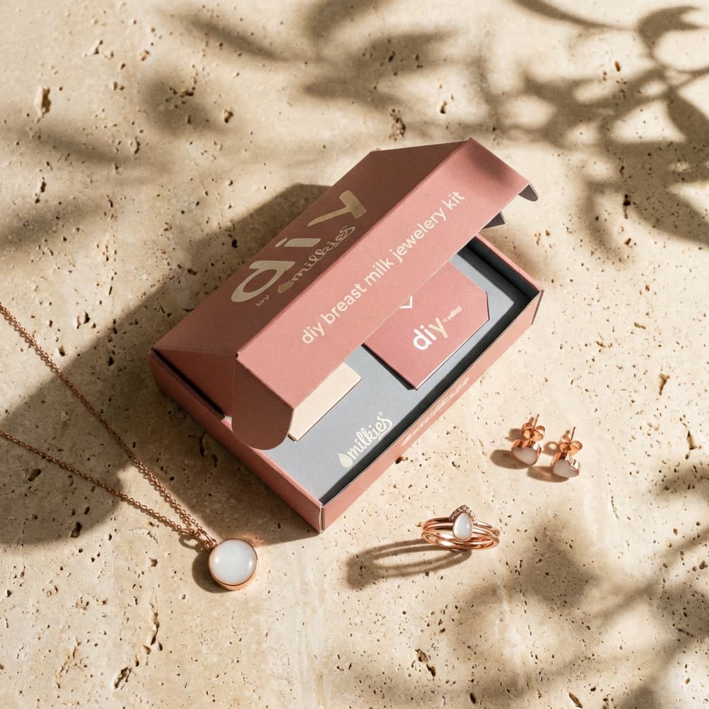 Boîte DIY by MILKIES ouverte présentant un kit création bijoux lait maternel avec accessoires et bijoux en finition or rose (collier, bague, boucles d’oreilles) ornés d’une pierre blanche, sur fond beige avec ombres de feuilles.