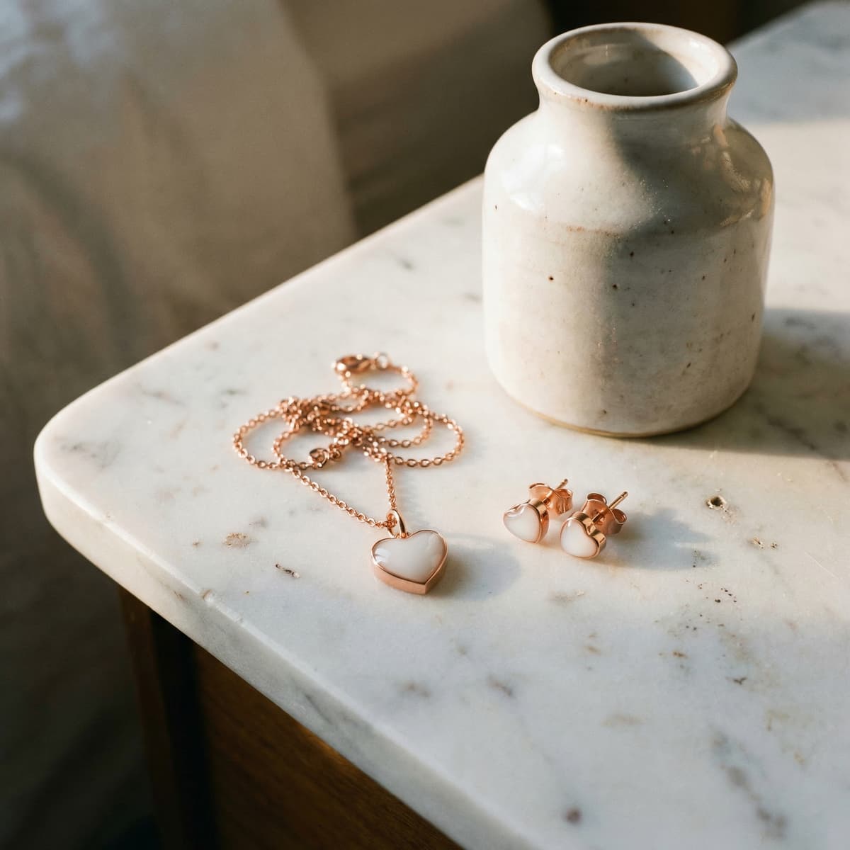 Collier et boucles d’oreilles en forme de cœur ton or rose posés sur une table en marbre près d’un vase, réalisés avec un kit diy bijoux lait maternel.