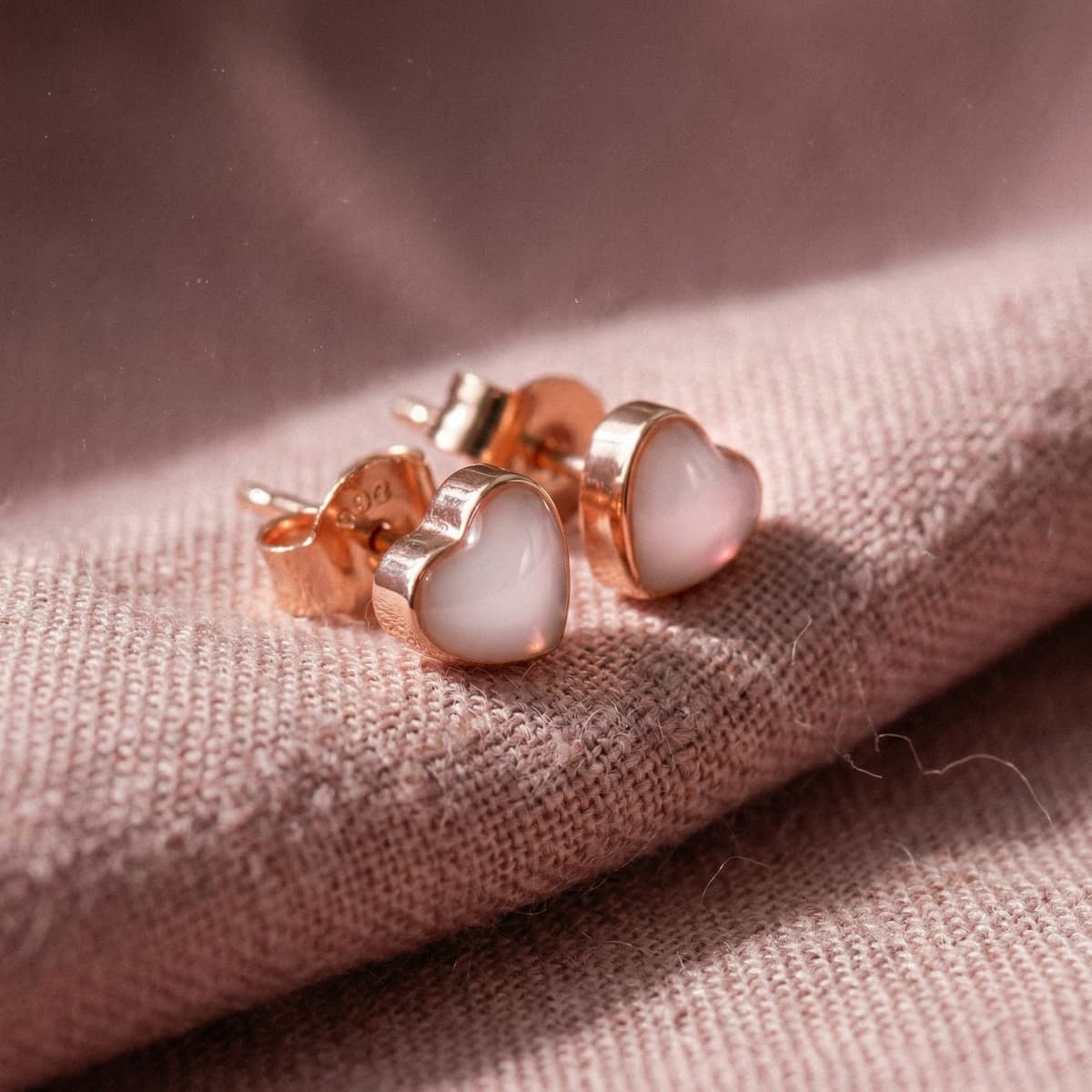 Gros plan de boucles d’oreilles cœur en métal doré rose, réalisées avec un kit fabrication boucles d'oreilles lait, posées sur un tissu rose texturé.
