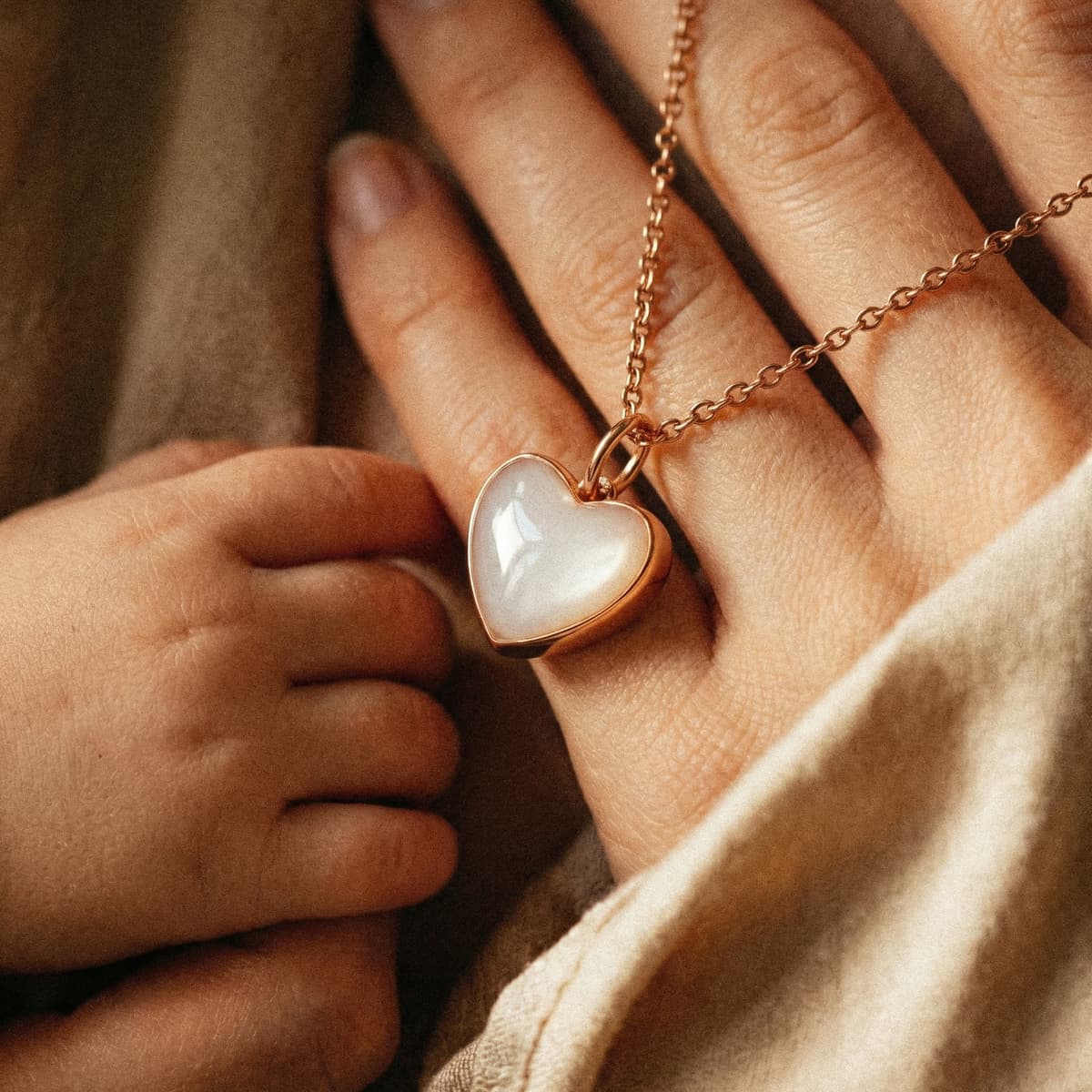 Gros plan d’un collier cœur en métal rose doré, symbole d’un kit fabrication collier lait maternel, posé sur la main d’une maman avec la main de son bébé sur une couverture douce.