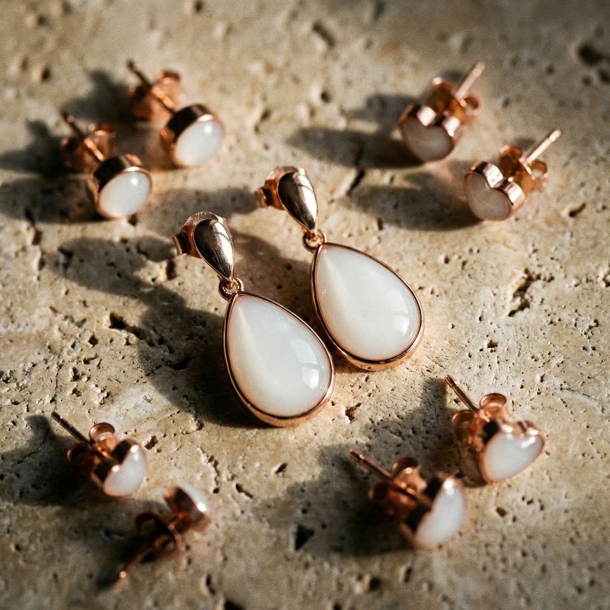 Collection de bijoux présentant une élégante paire de boucles d'oreilles lait maternel en forme de goutte, sertie en rose doré avec pierre blanche laiteuse, posée sur un fond de pierre texturée avec petits clous assortis.