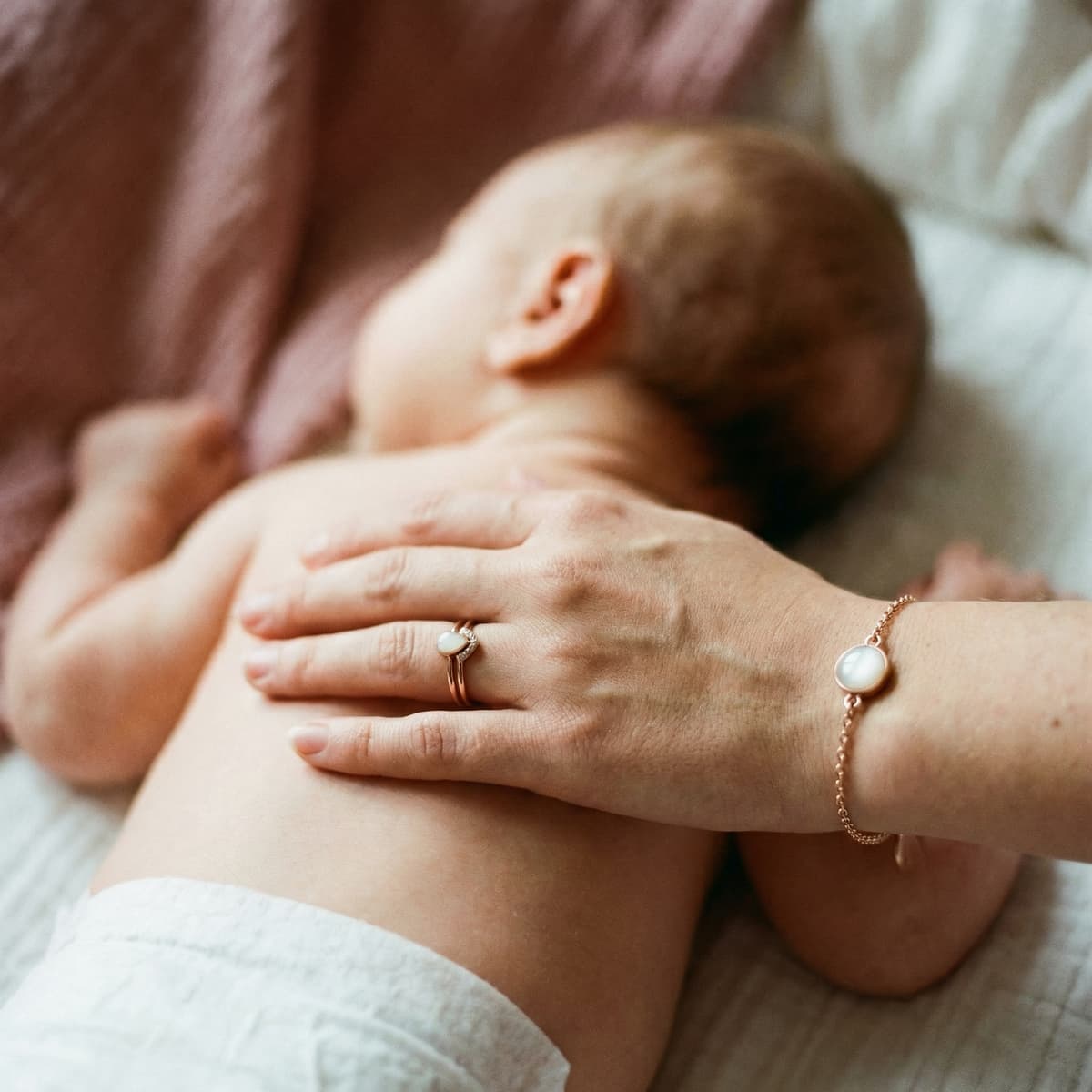 Main d’une maman caressant un bébé allongé, avec bague et bracelet dorés ornés d’une pierre lactée nacrée, évoquant un souvenir maternel précieux.